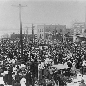 Large mixed crowd in town street with people on rooftops and on train cars in the background