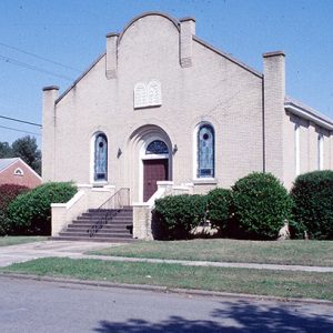 Single-story synagogue building with bushes on street corner with brick church in background