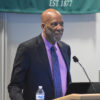 African-American man with beard in suit and tie speaking into microphone at lectern