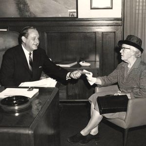 White man in suit and tie sitting at desk handing something to elderly white woman with glasses and hat in chair