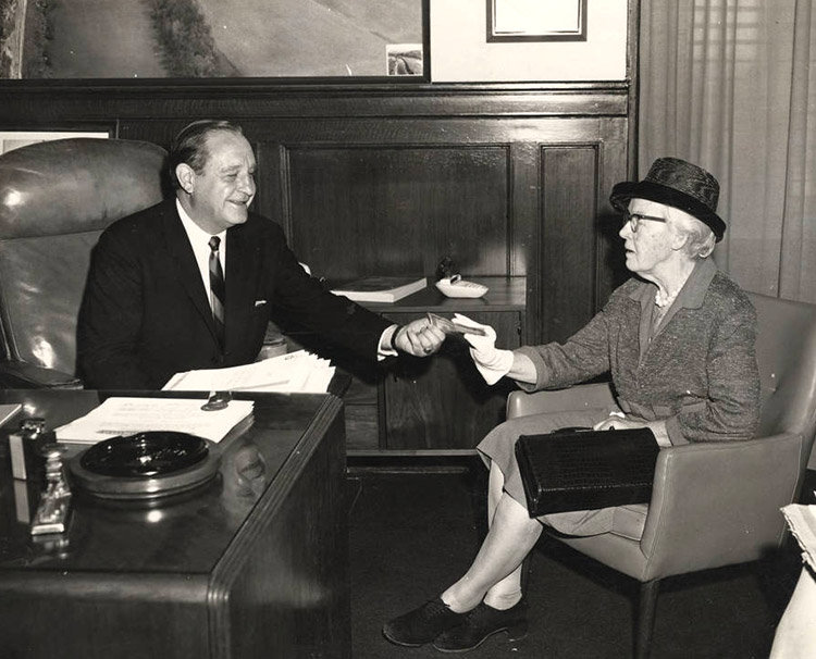 White man in suit and tie sitting at desk handing something to elderly white woman with glasses and hat in chair