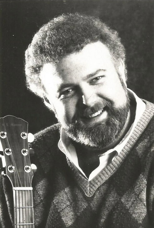 White man smiling in beard and sweater with guitar