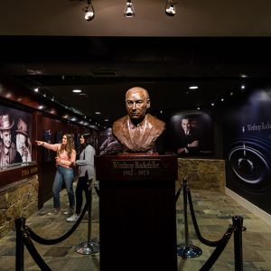 Two young women standing behind a bust of white man looking at photographs in museum exhibit room