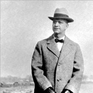 White man in long coat and fedora hat holding a suitcase