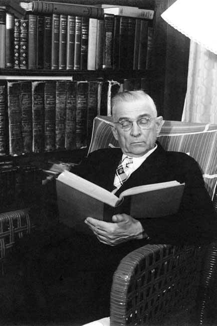 Portrait of a man in formal attire reading in a wicker chair next to a bookcase.