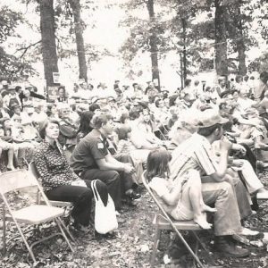 Crowd of white men women and children sitting in folding chairs under trees at political rally