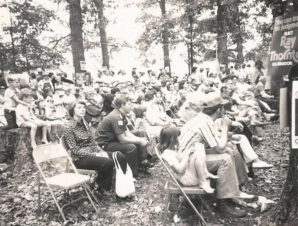 Crowd of white men women and children sitting in folding chairs under trees at political rally