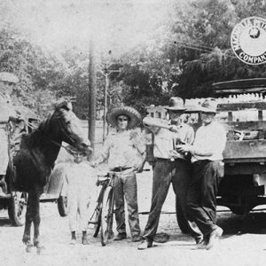 White boys on horse and standing beside horse with man leaning on bicycle and two white men standing behind a truck filled with boxes of bottles