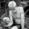 Old white man sitting on large stones shirtless in shorts holding a rock in his right hand