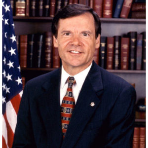 Smiling white man posing in suit and tie with flag and bookshelf in background