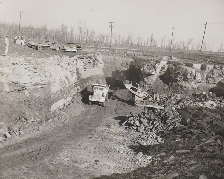 White workers and trucks parked above open pit with bull dozer loading dump truck at the bottom of it