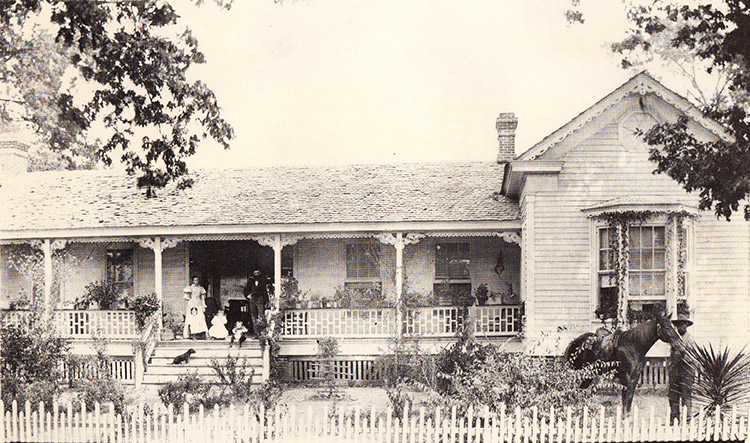 White family on steps of single-story house with covered porch and fence and man with horse nearby
