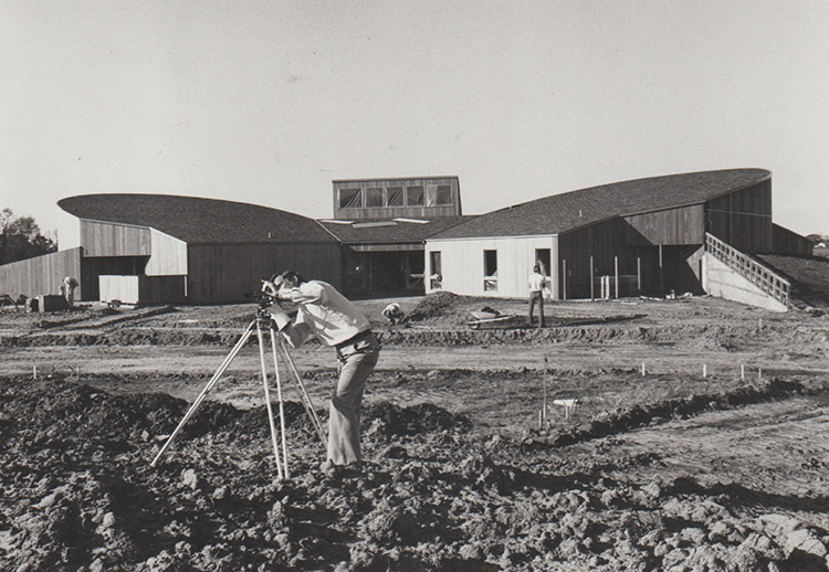White man with surveying equipment standing in field with white men and building with rounded wings and skylight behind them