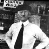 white man in beret and tie standing before shelves of various goods