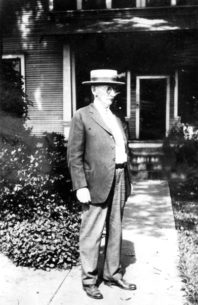 white man in straw hat stands outside wooden house