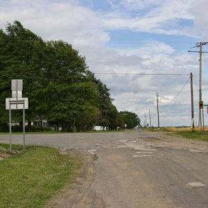 Damaged rural roads with road sign and house under trees on its left side under power lines
