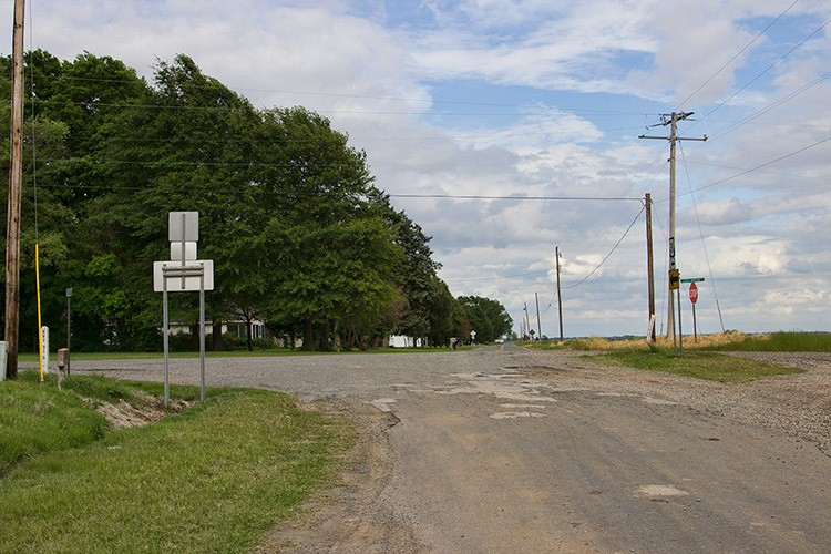 Damaged rural roads with road sign and house under trees on its left side under power lines
