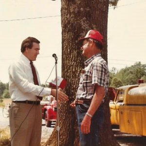 White man in shirt and tie at microphone under a tree shaking hands with white man in hat plaid shirt and jeans