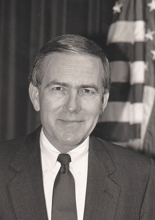 White man smiling in suit and tie with flag behind him