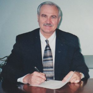 Older white man with mustache smiling in suit and tie while at desk with paper and pen in hands