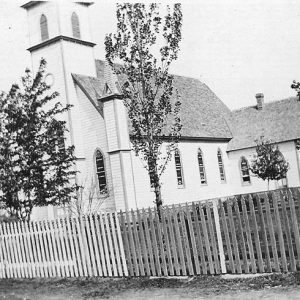 High pitched wood frame church with spires behind picket fence along road