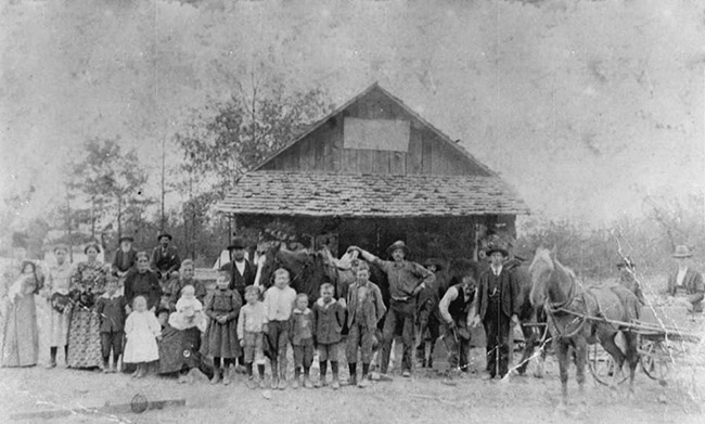 Group of white men women children and horse drawn wagon outside single-story building