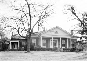 Single-story brick house with covered porch entrance with columns with trees in front