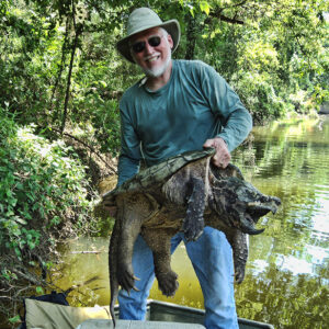 White man wearing sunglasses hat and green shirt holding an alligator snapping turtle in boat