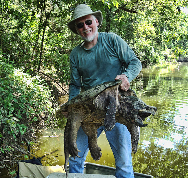 White man wearing sunglasses hat and green shirt holding an alligator snapping turtle in boat