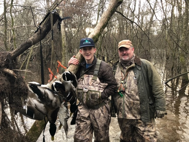 Young white teenage boy and older white man in hunting gear in swamp with ducks hanging from a tree