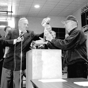 Old white man handing a football trophy to another old white man in school gymnasium