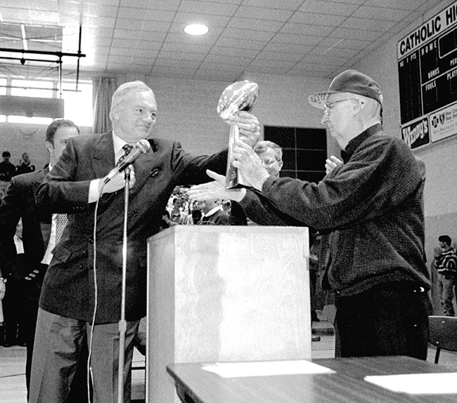 Old white man handing a football trophy to another old white man in school gymnasium