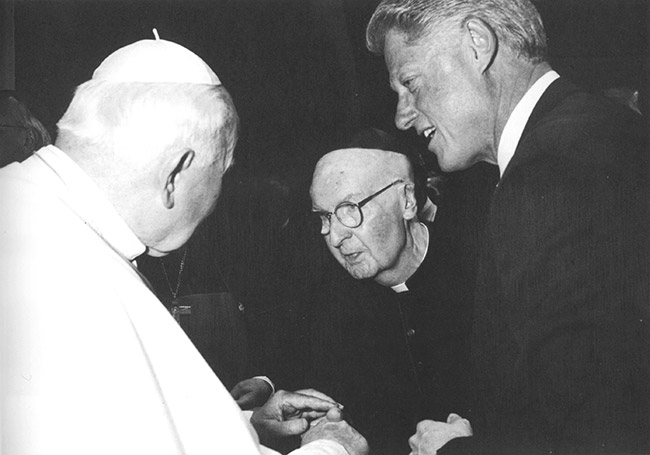 White man in suit talking to old white man in robes and old white man in black clerical outfit with white collar