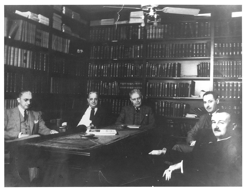 Five white men in suits in library seated around a table