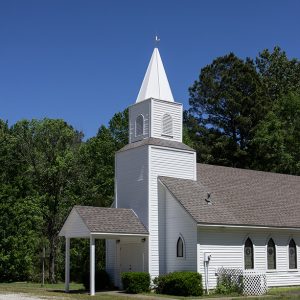 Single-story church building with white siding steeple tower and covered entrance on gravel driveway