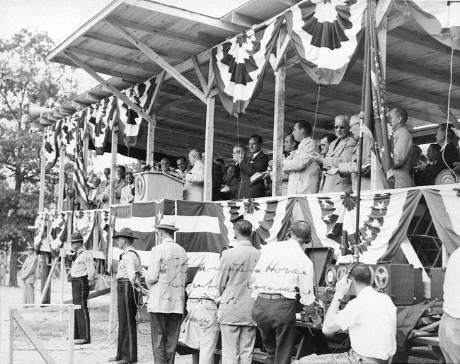 White man at lectern speaking to white crowd from a covered stage decorated with bunting