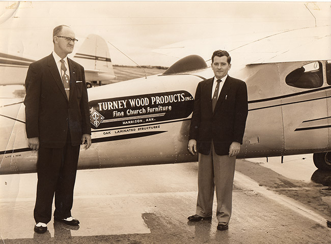 Two white men in suit and tie posing with "Turney Wood Products" logo on airplane