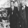 Two white men in suits with dog and house behind them