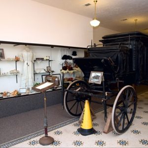 Black stagecoach with wooden wheels in museum gallery next to glass display case