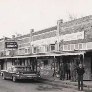 Group of men standing on sidewalk in front of row of storefronts