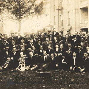 Group of young white men in suits posing with skeleton outside multistory buildings
