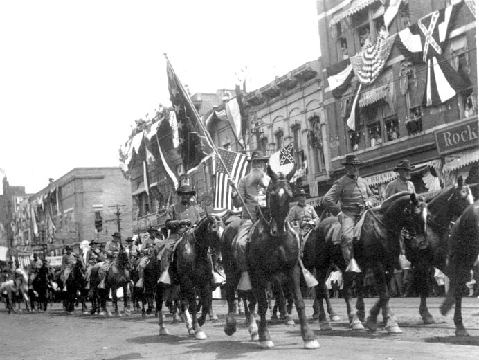 White men in Confederate uniforms and hats holding Confederate flags ride on horseback in a parade