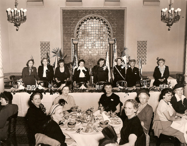 Group of older white women in formal clothing posing at tables in dining hall