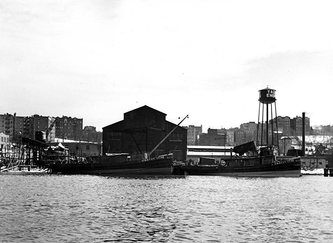 Boats moored at shipping depot with water tower and buildings in the background