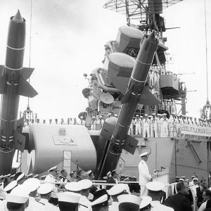White man in Naval uniform speaking to sailors on deck of naval warship with missiles pointed towards the sky