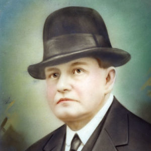White man with black hat in black suit and blue-green background