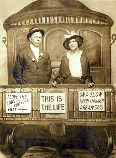White man and woman in suit and dress posing with fake train car set featuring signs such as "I love the cows and chickens but" and "This is the life" and "On a slow train through Arkansas."