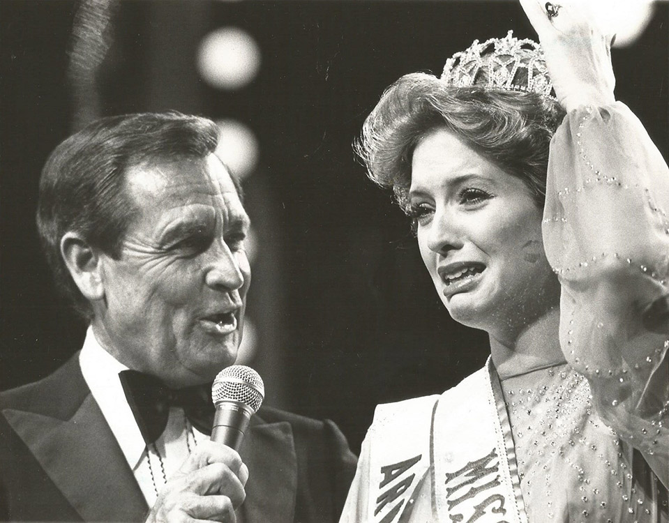 Older white man in suit and bow tie talking into microphone next to young woman in dress sash and tiara