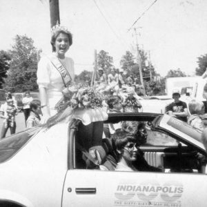 White woman in dress and tiara riding in car in parade