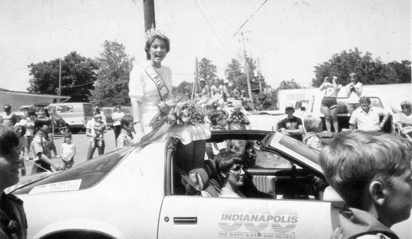 White woman in dress and tiara riding in car in parade